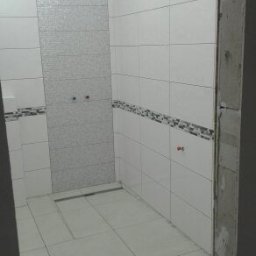 KD EXPERT Konrad Drzazga - Wnętrze łazienki w trakcie wykańczania, widoczne białe płytki ceramiczne na ścianach z dekoracyjnym pasem płytek mozaikowych, podłoga wyłożona jasnymi płytkami, odpływ liniowy prysznicowy...