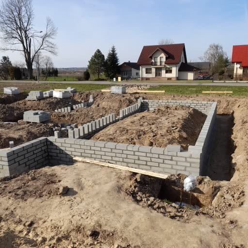 Wykop pod fundamenty domu z częściowo wymurowanymi ścianami z szarych bloczków betonowych, widoczne ułożone bloczki na ziemi i drewniana deska oparta o mur.