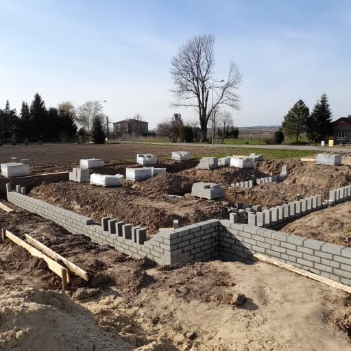 Wznoszenie muru fundamentowego z bloczków betonowych na placu budowy, widoczne wystające pręty zbrojeniowe, w tle przygotowane bloczki i teren pod budowę.