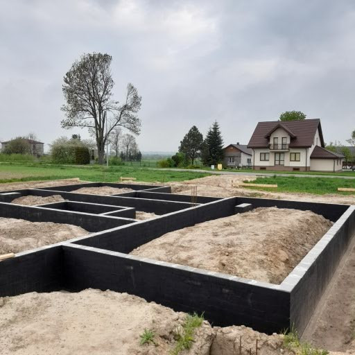 Przygotowane fundamenty domu jednorodzinnego z hydroizolacją, widoczne zbrojenia i szalunki, w tle dom w budowie i drzewo, pochmurne niebo.