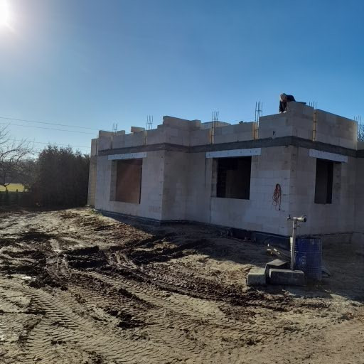 Budowa domu z betonu komórkowego w trakcie realizacji, widoczny pracownik na dachu, zbrojenie, okna bez szyb, teren wokół z błotem i śladami opon.