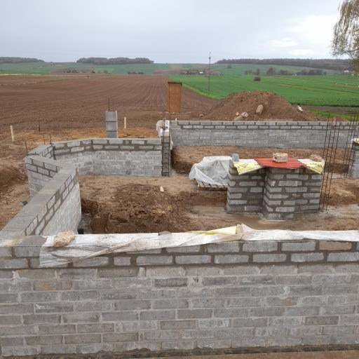 Fundamenty domu w trakcie budowy, widoczne bloczki betonowe i zbrojenie, w tle krajobraz polny pod pochmurnym niebem.