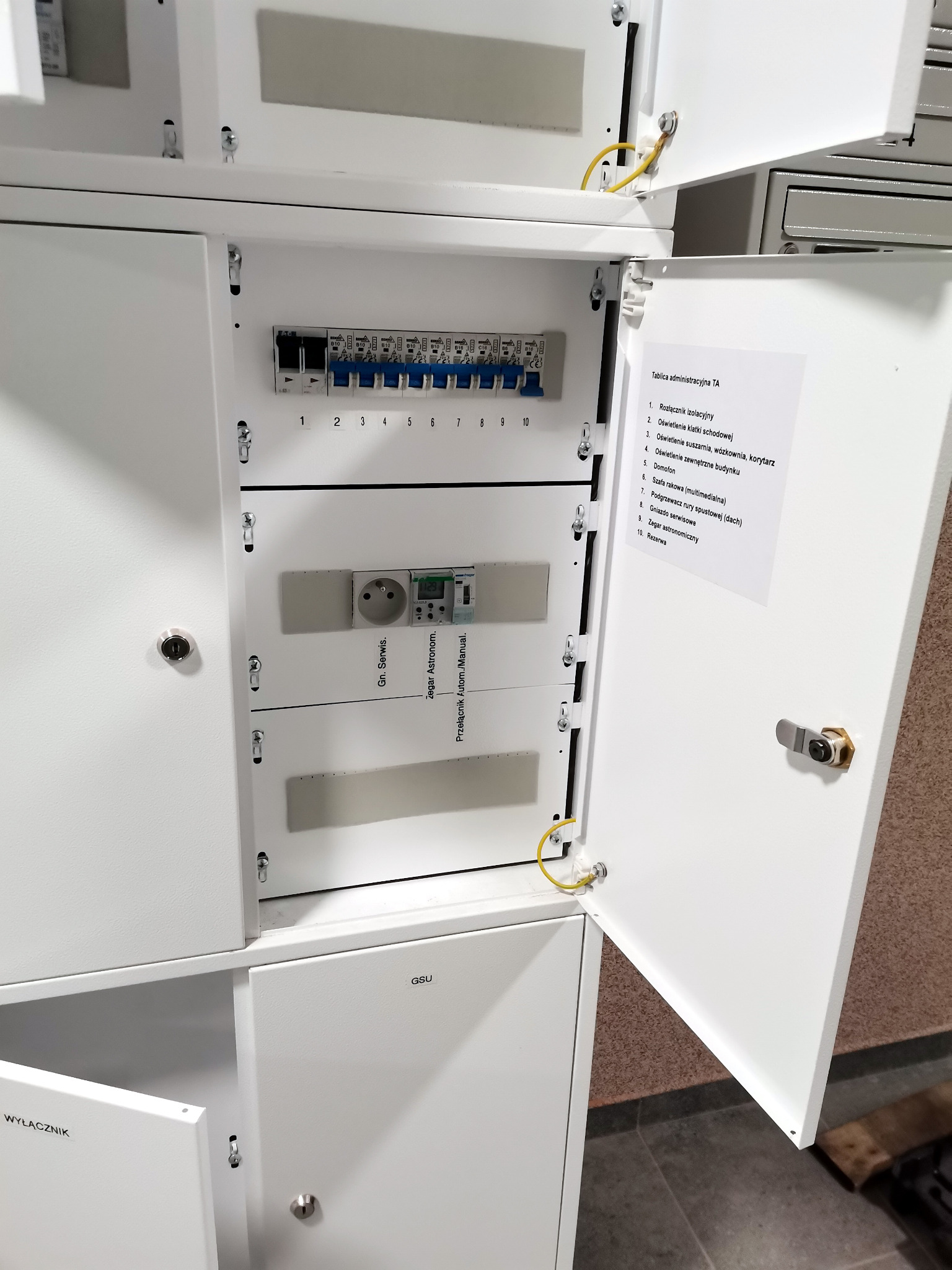 Otwarta szafa rozdzielcza z widocznymi bezpiecznikami, gniazdem serwisowym i zegarem astronomicznym, z żółtym przewodem uziemiającym oraz naklejką z opisem funkcji poszczególnych elementów.