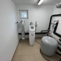 Biały zmiękczacz wody Helix 25L w pomieszczeniu technicznym z widocznymi rurami instalacji wodnej i zbiornikiem wyrównawczym, oświetlonym jarzeniówką.