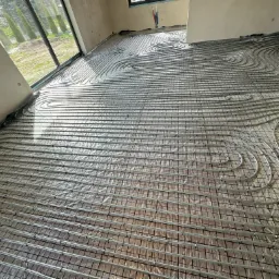 Rozłożone rury ogrzewania podłogowego na siatce montażowej w surowym wnętrzu, widoczne okno z widokiem na ogród.