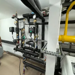 Węzeł cieplny z czarnymi rurami izolowanymi termicznie, zaworami, manometrami, sterownikami i żółtą rurą gazową, zamontowany w pomieszczeniu technicznym z wykończoną podłogą z płytek.