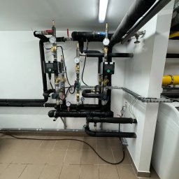 Zakład Usługowy Instalacje C.O., Wod.-Kan. i Gaz Usługi Ślusarskie Leszek Zdrzałka - Złożona instalacja rur, zaworów i manometrów w kotłowni, z czarnymi izolowanymi rurami i elementami sterującymi Wilo na białej ścianie, widoczne płytki podłogowe i fragment bojlera.