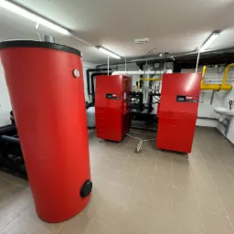 Kotłownia z czerwonym zasobnikiem ciepłej wody i dwoma czerwonymi kotłami gazowymi marki Hoval, widoczne rury instalacji grzewczej i gazowej oraz umywalka.