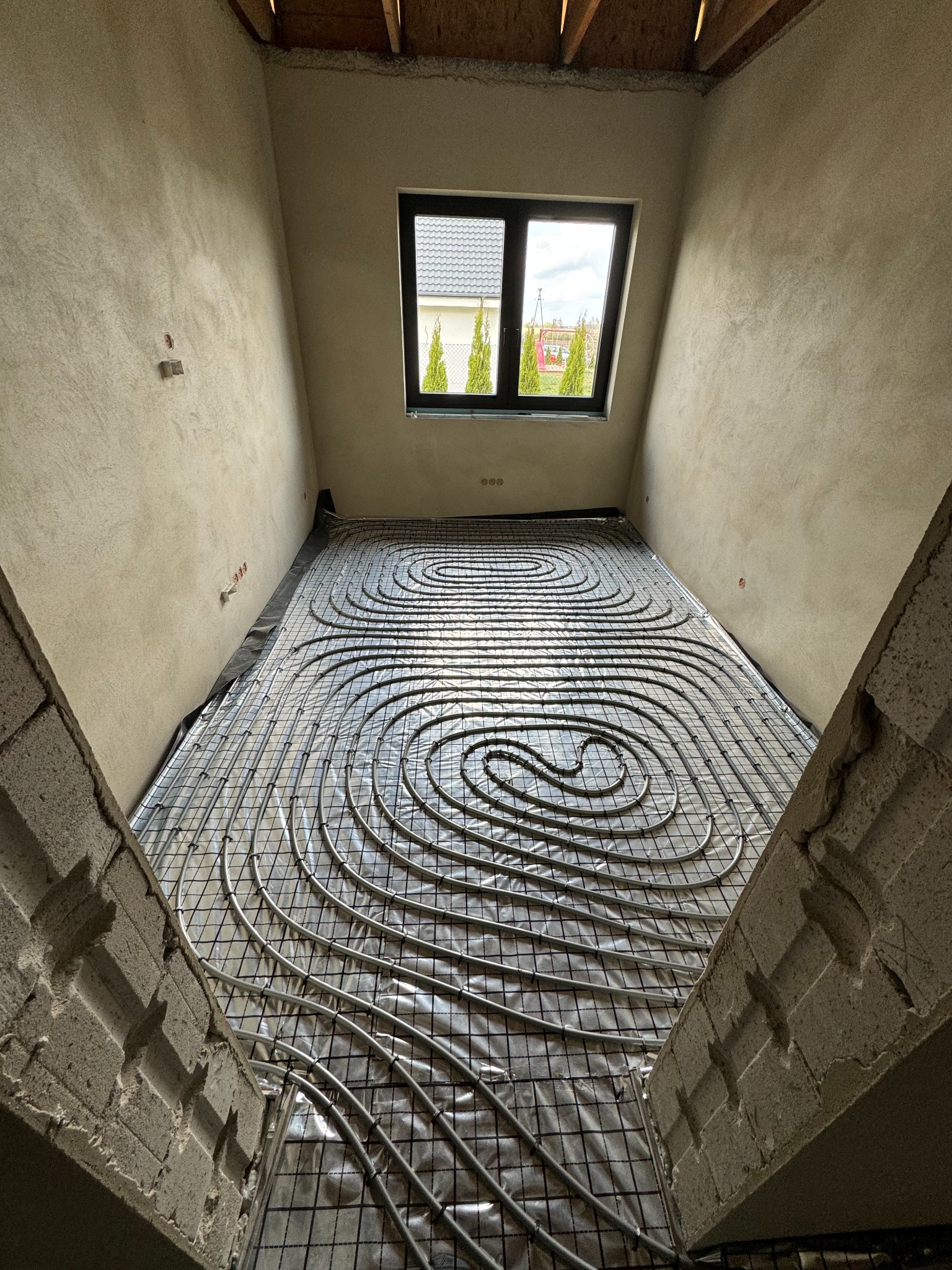 Widok z góry na rozłożone rury ogrzewania podłogowego na folii odblaskowej, zamocowane do siatki metalowej w surowym pomieszczeniu z oknem.