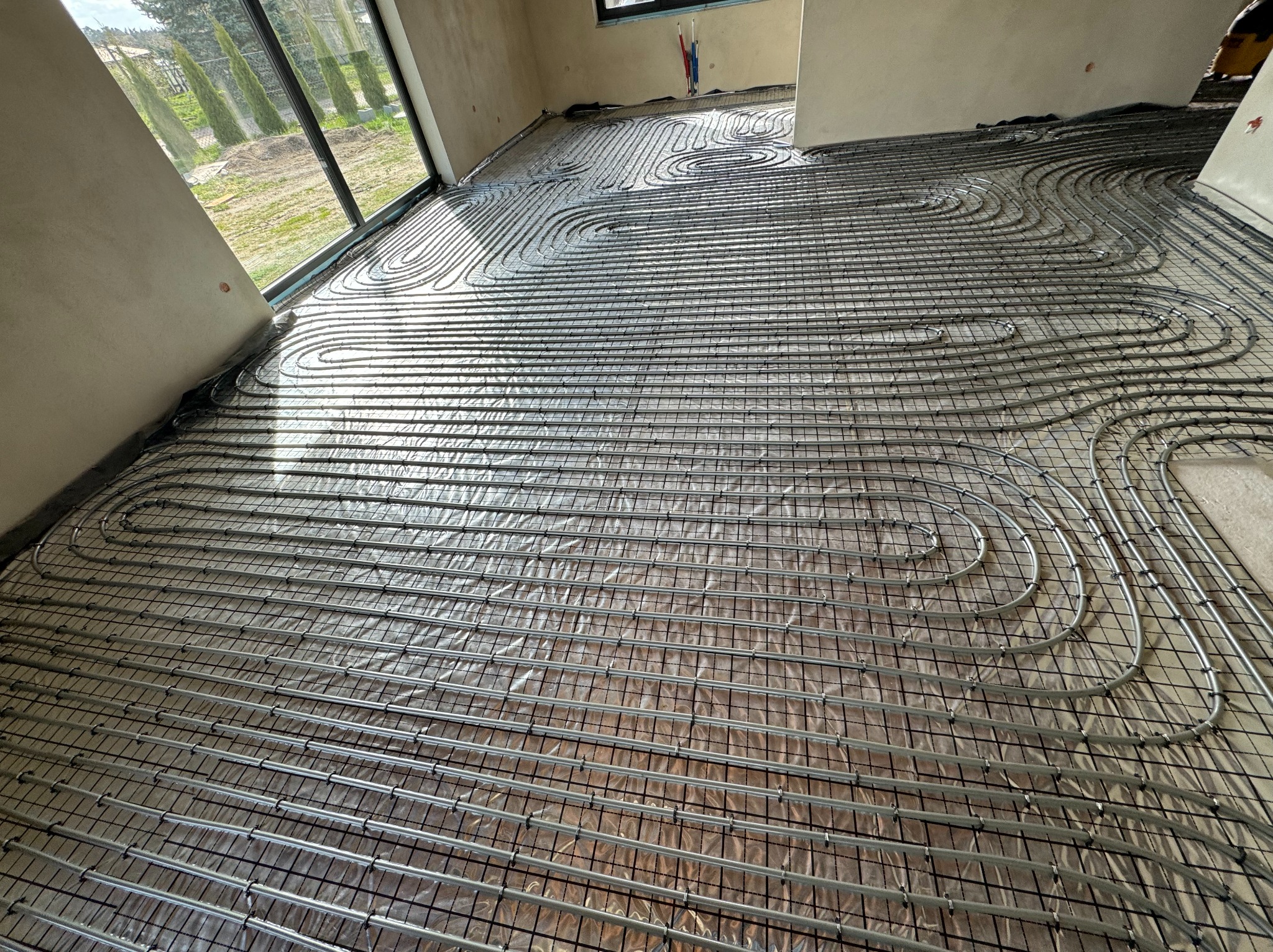 Rozłożone rury ogrzewania podłogowego na folii izolacyjnej z siatką w surowym wnętrzu, widok z góry pod kątem.