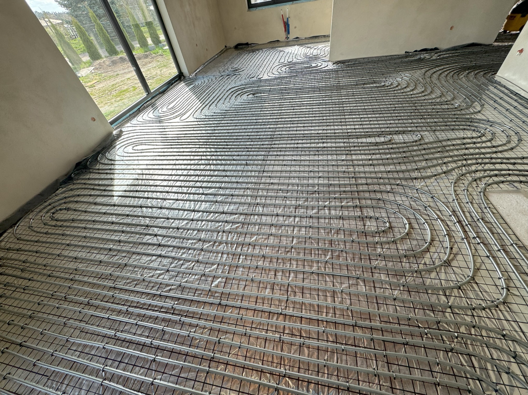 Rozłożone rury ogrzewania podłogowego na siatce montażowej w surowym wnętrzu, widoczne okno z widokiem na ogród.