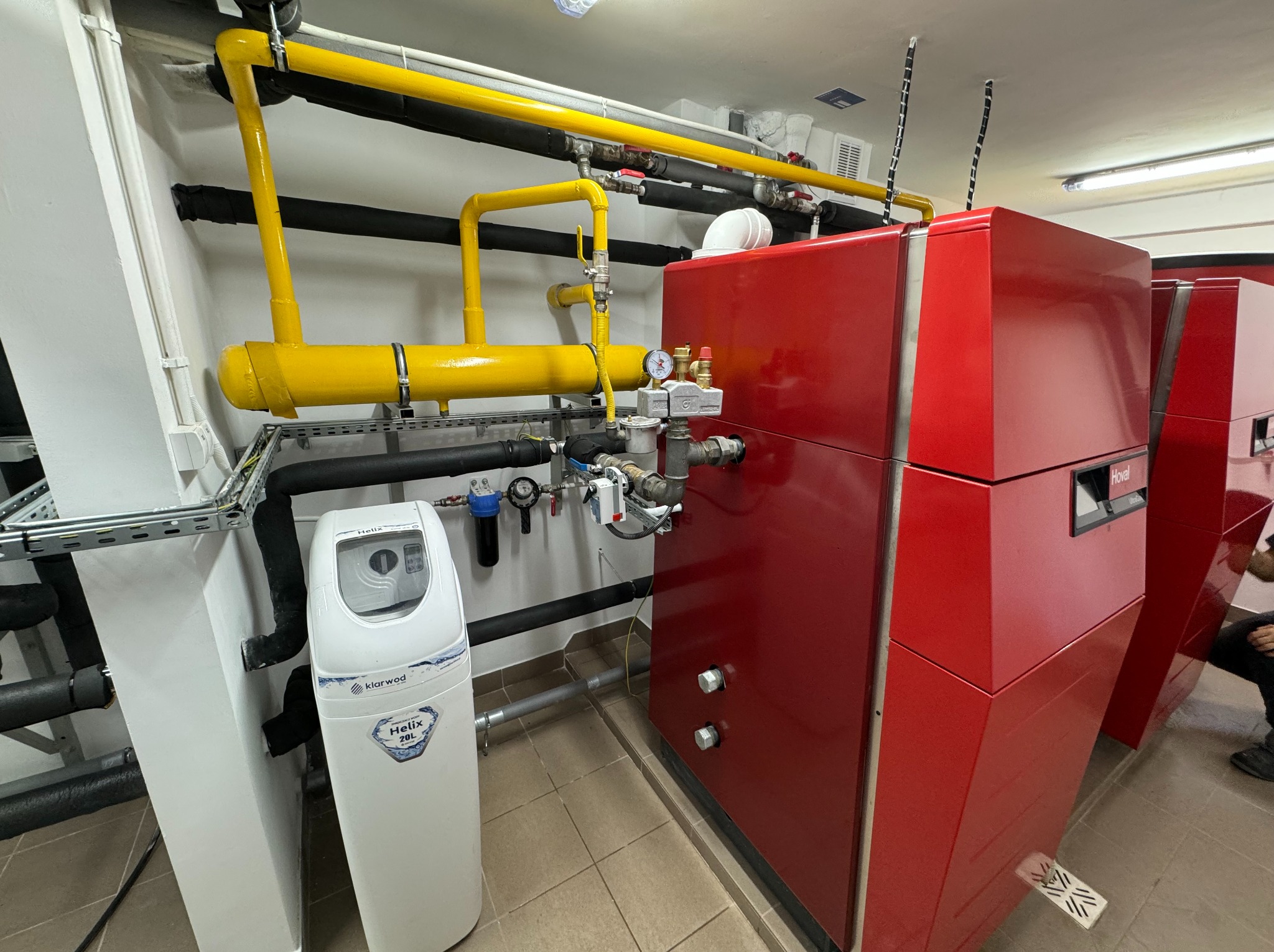 Czerwony piec grzewczy marki Hoval z żółtymi rurami gazowymi i białym zmiękczaczem wody Klarwod Helix 20L w kotłowni.