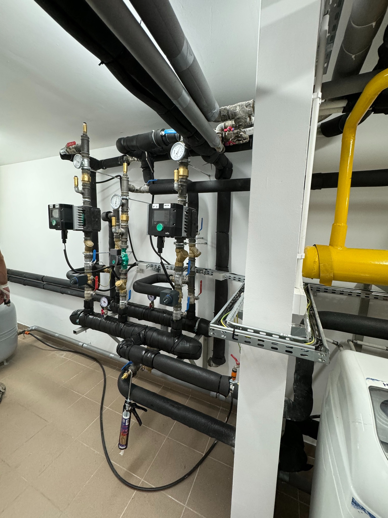Węzeł cieplny z czarnymi rurami izolowanymi termicznie, zaworami, manometrami, sterownikami i żółtą rurą gazową, zamontowany w pomieszczeniu technicznym z wykończoną podłogą z płytek.