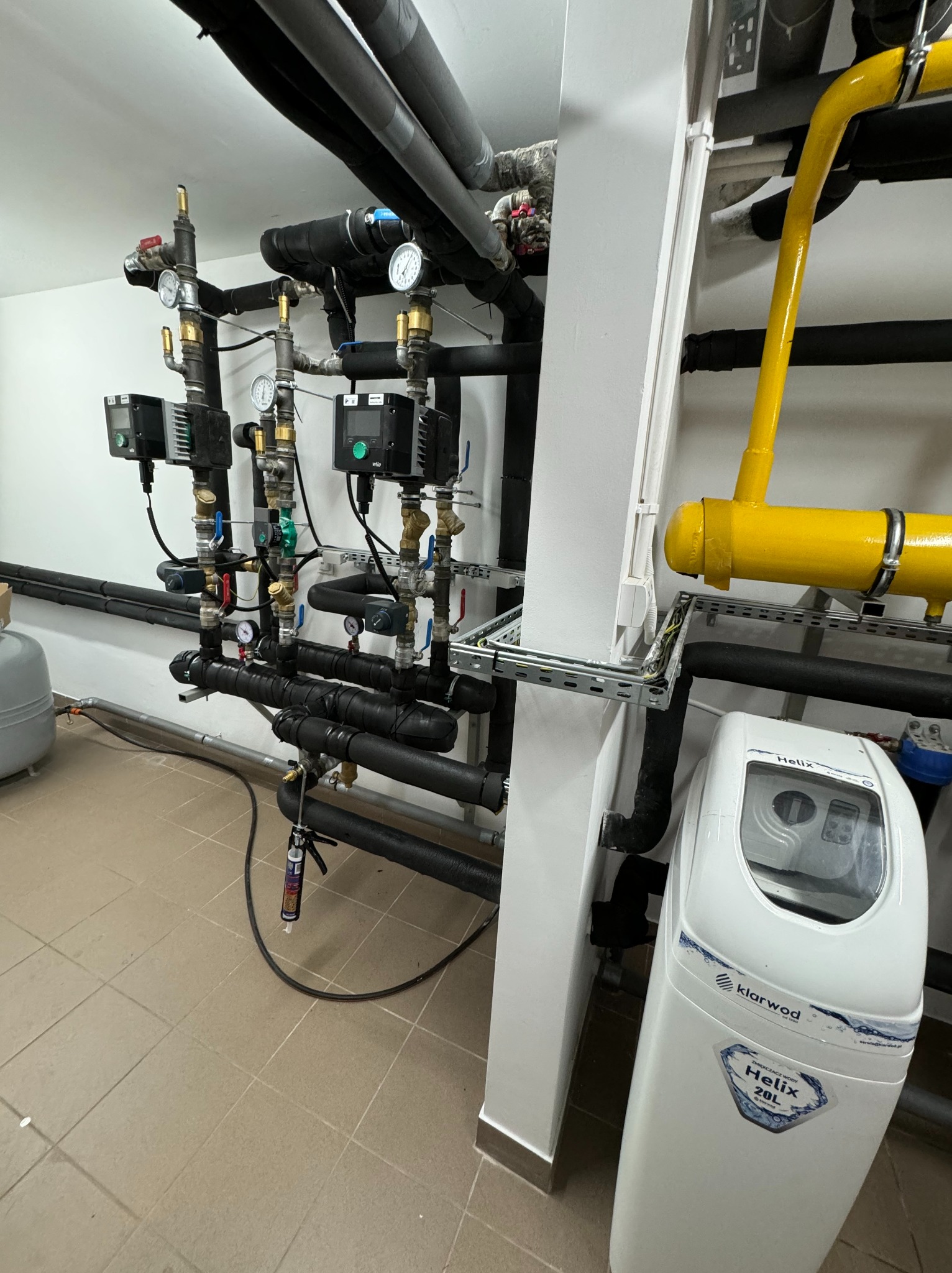Węzeł cieplny w kotłowni: czarne rury izolacyjne, manometry, pompy obiegowe Wilo, żółta rura gazowa, zmiękczacz wody Klarwod Helix 20L, aplikator silikonu na podłodze.