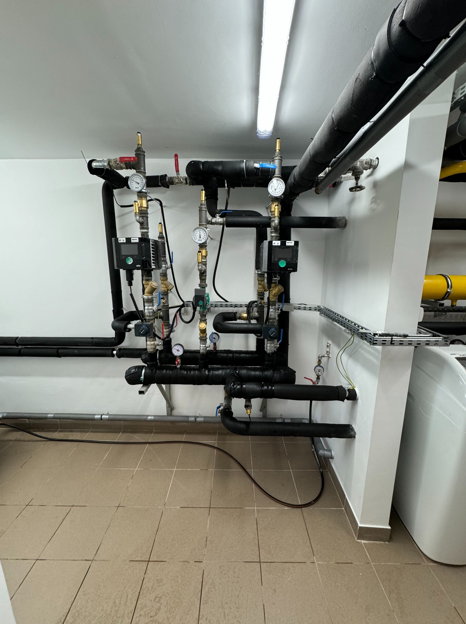 Złożona instalacja rur, zaworów i manometrów w kotłowni, z czarnymi izolowanymi rurami i elementami sterującymi Wilo na białej ścianie, widoczne płytki podłogowe i fragment bojlera.