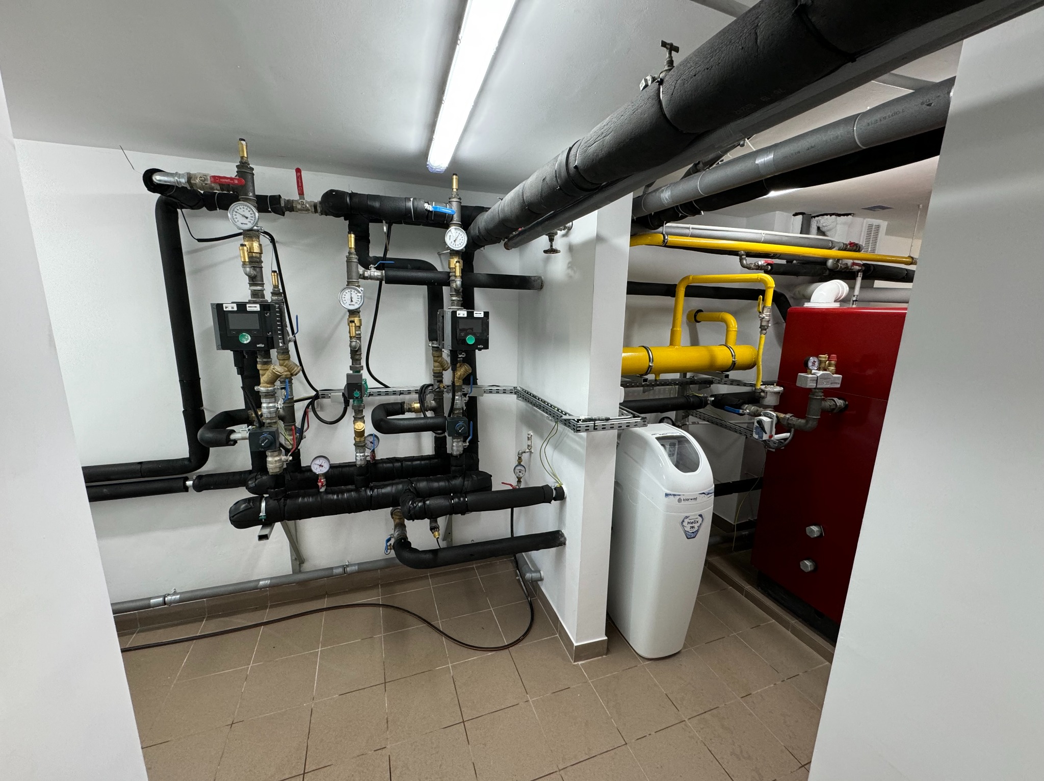 Węzeł instalacji hydraulicznej i grzewczej z czarnymi rurami izolowanymi termicznie, manometrami, zaworami, sterownikami, czerwonym zasobnikiem ciepłej wody i białą stacją uzdatniania wody Klarwod...