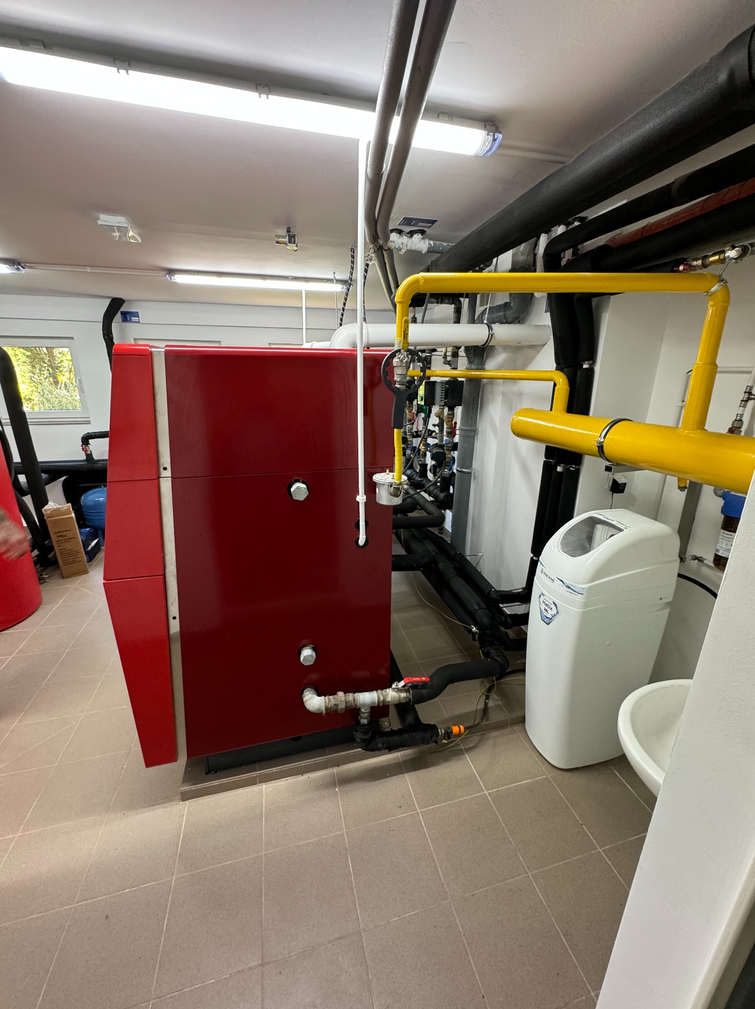Czerwony piec grzewczy z podłączonymi rurami w kotłowni, widoczny zmiękczacz wody i instalacja gazowa z żółtymi rurami.