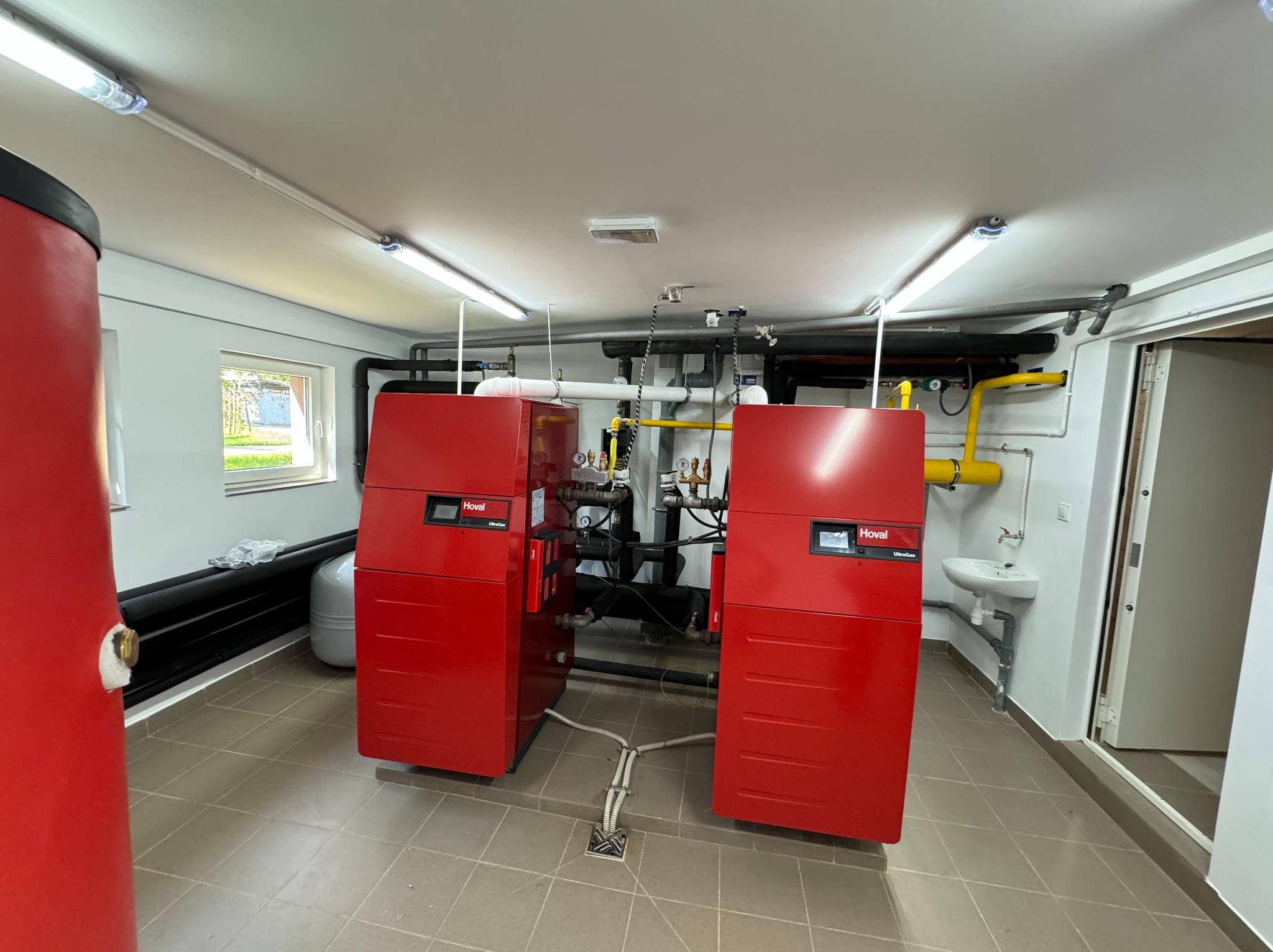 Dwa czerwone piece gazowe marki Hoval UltraGas w kotłowni w Wągrowcu, widoczne rury i instalacje, umywalka oraz okno z widokiem na zewnątrz.