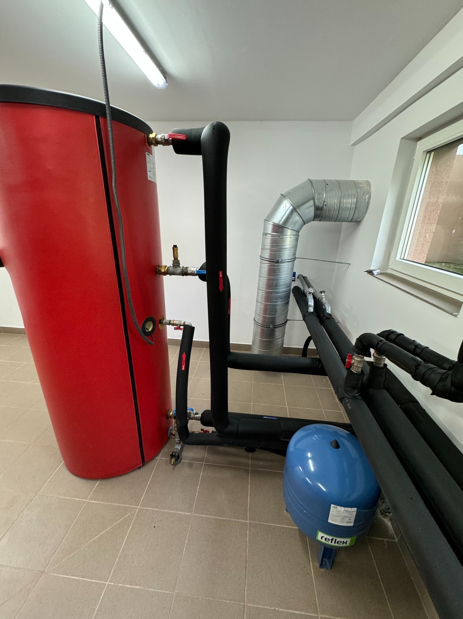 Czerwony zasobnik ciepłej wody użytkowej z czarną izolacją, podłączony do czarnych rur z izolacją, widoczny niebieski zbiornik wyrównawczy Reflex i fragment metalowego przewodu wentylacyjnego...