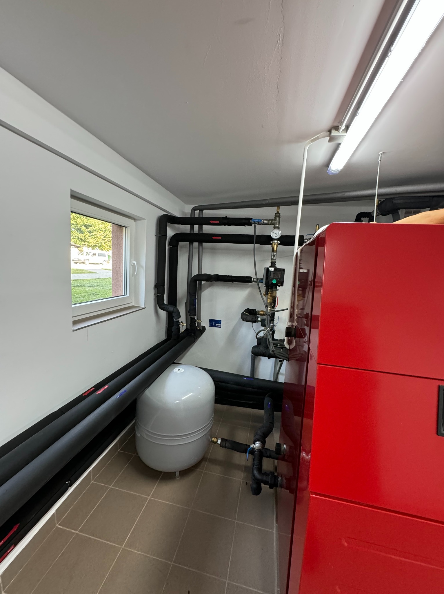 Czerwony piec grzewczy z podłączonymi rurami w izolacji, szary zbiornik wyrównawczy, instalacja i okno z widokiem na zewnątrz w jasnym pomieszczeniu.