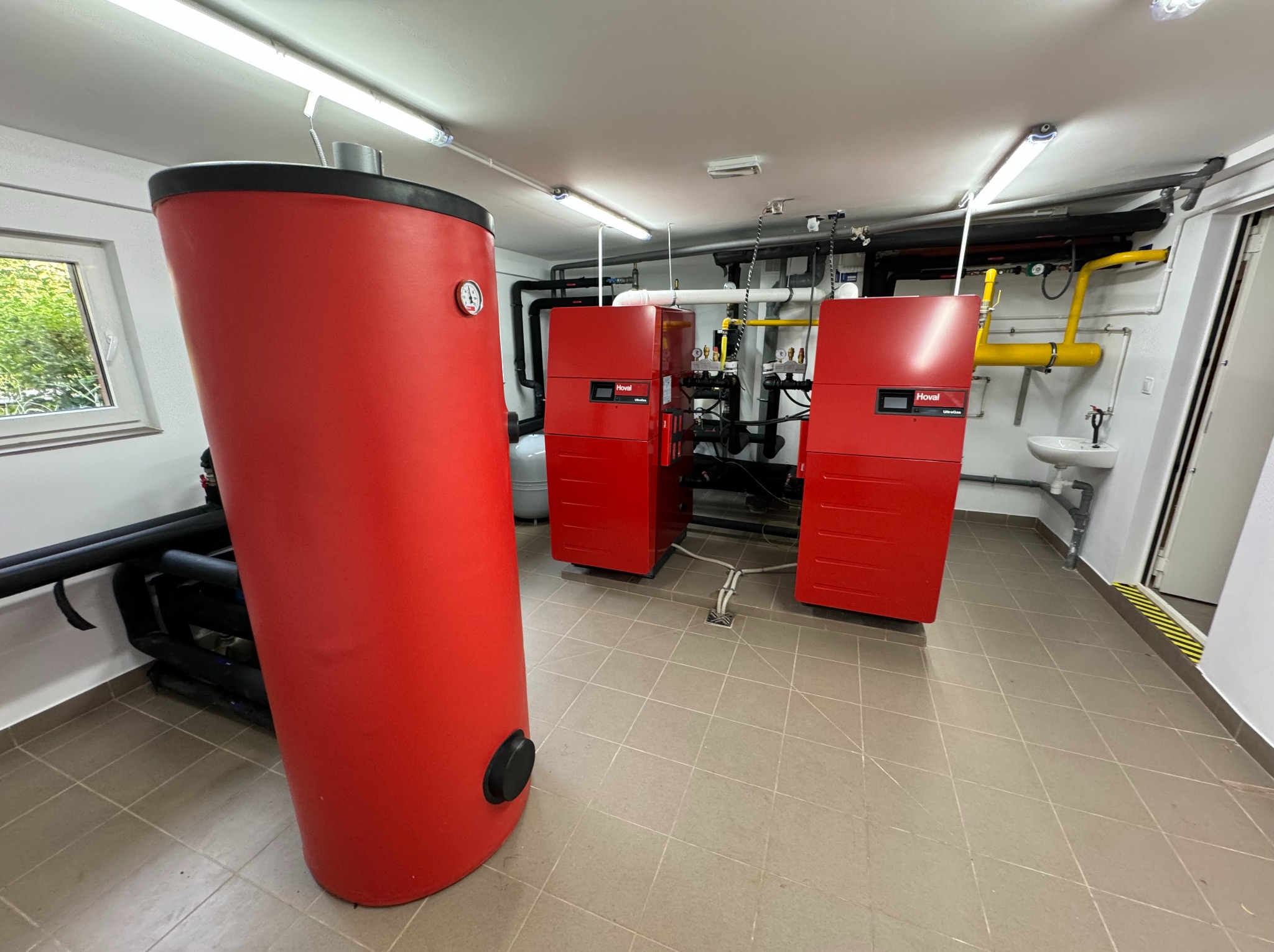 Kotłownia z czerwonym zasobnikiem ciepłej wody i dwoma czerwonymi kotłami gazowymi marki Hoval, widoczne rury instalacji grzewczej i gazowej oraz umywalka.