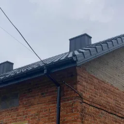 Ciemnoszara dachówka ceramiczna na dachu murowanego domu, rynna spustowa z tworzywa sztucznego, widoczne kominy wentylacyjne.