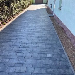 BRUK-BAU ZBOROWSKI BARTŁOMIEJ ZBOROWSKI - Długa, prosta droga z szarej kostki brukowej prowadząca do zielonego garażu, z żywopłotem po lewej stronie i ścianą budynku po prawej, z widocznym fragmentem rynny.