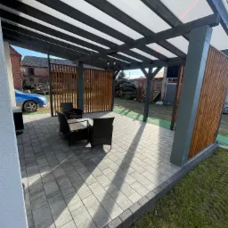 Zadaszony taras z szarych płyt betonowych, obrzeża z ciemnej kostki, meble ogrodowe z rattanu, drewniane panele osłonowe, w tle zabudowania gospodarcze i stos drewna.