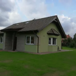 Niski, parterowy dom z zieloną elewacją i brązowym dachem, fragment ściany wykończony kamieniem elewacyjnym, schludny trawnik.