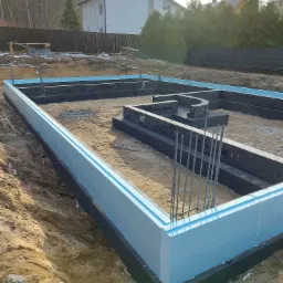 Fundamenty domu z izolacją termiczną w postaci niebieskiego styropianu, częściowo zasypane ziemią, z wystającymi prętami zbrojeniowymi, w tle niezabudowana działka z drzewami i domem.