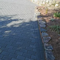 Ułożona z szarej kostki brukowej ścieżka zakręca obok kamiennego murku rabaty z roślinami.