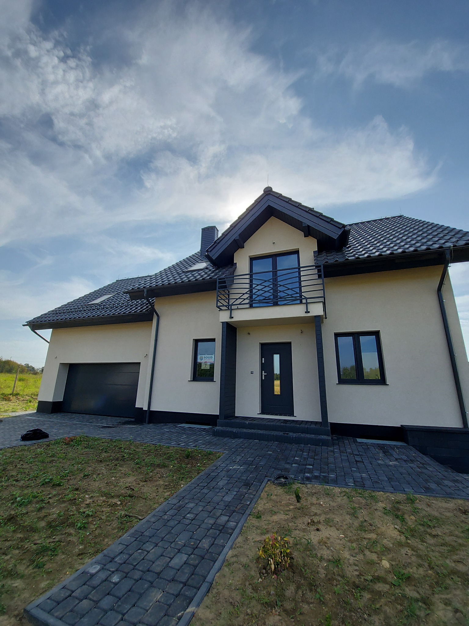 Nowoczesny, dwukondygnacyjny dom murowany z czarną dachówką, balkonem z metalową balustradą i garażem, widoczny z perspektywy niskiego kąta, z częściowo zagospodarowanym terenem przed budynkiem.