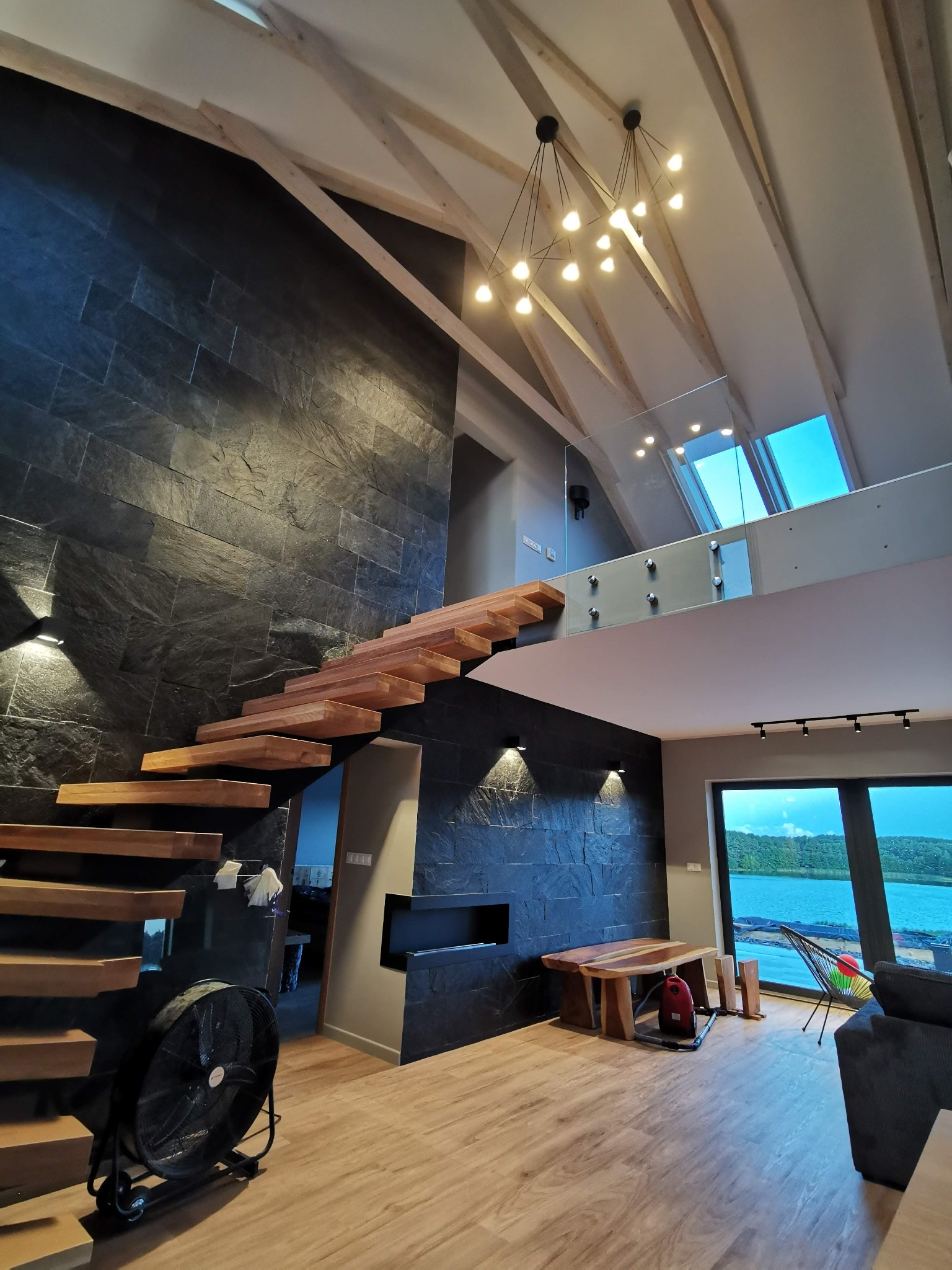 Wnętrze domu z drewnianymi schodami wspornikowymi, ścianą wykończoną czarnym kamieniem łupkowym, widokiem na jezioro z dużego okna i nowoczesnym oświetleniem sufitowym.