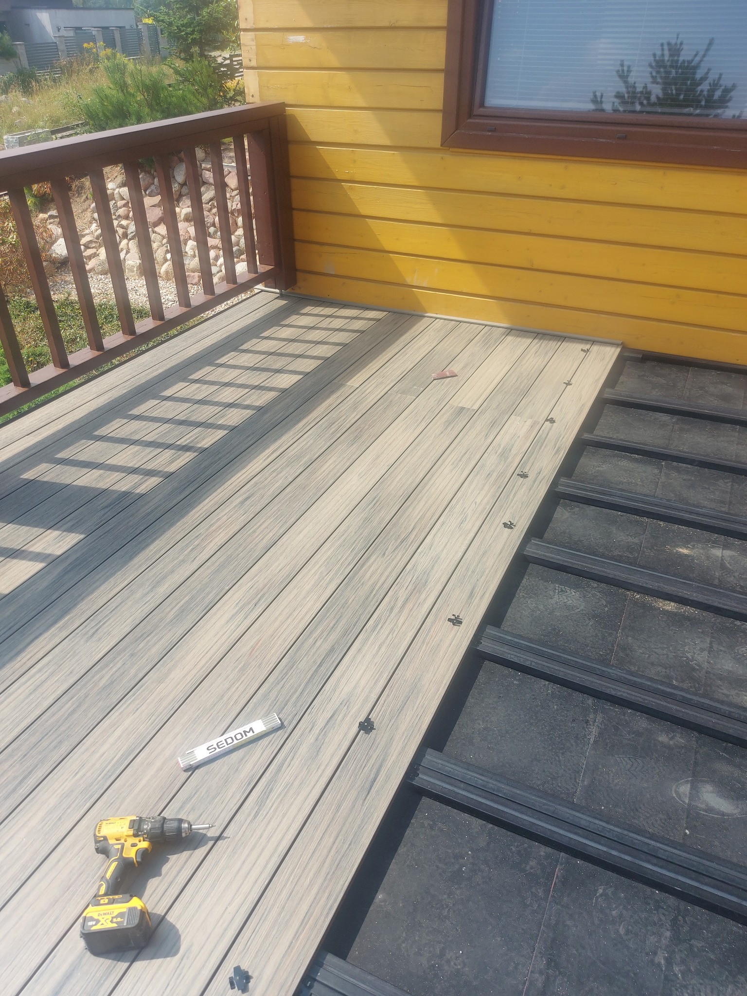 Montaż deski kompozytowej na tarasie domu z żółtą elewacją, widoczny fragment drewnianej balustrady, wkrętarka DeWalt i miarka Sedom na ułożonej części podłogi.