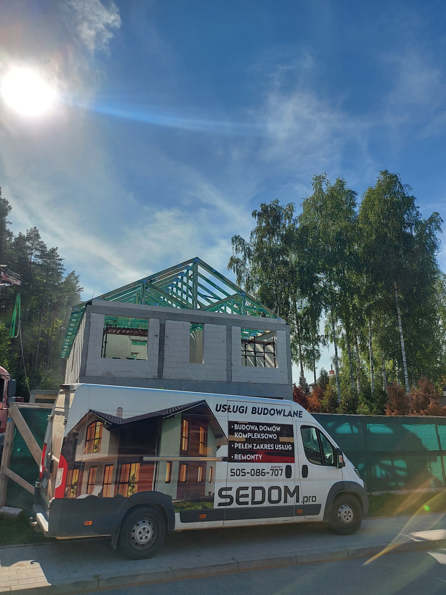 Budowa domu w stanie surowym otwartym z widoczną konstrukcją dachu, na pierwszym planie samochód firmowy z grafiką przedstawiającą dom.