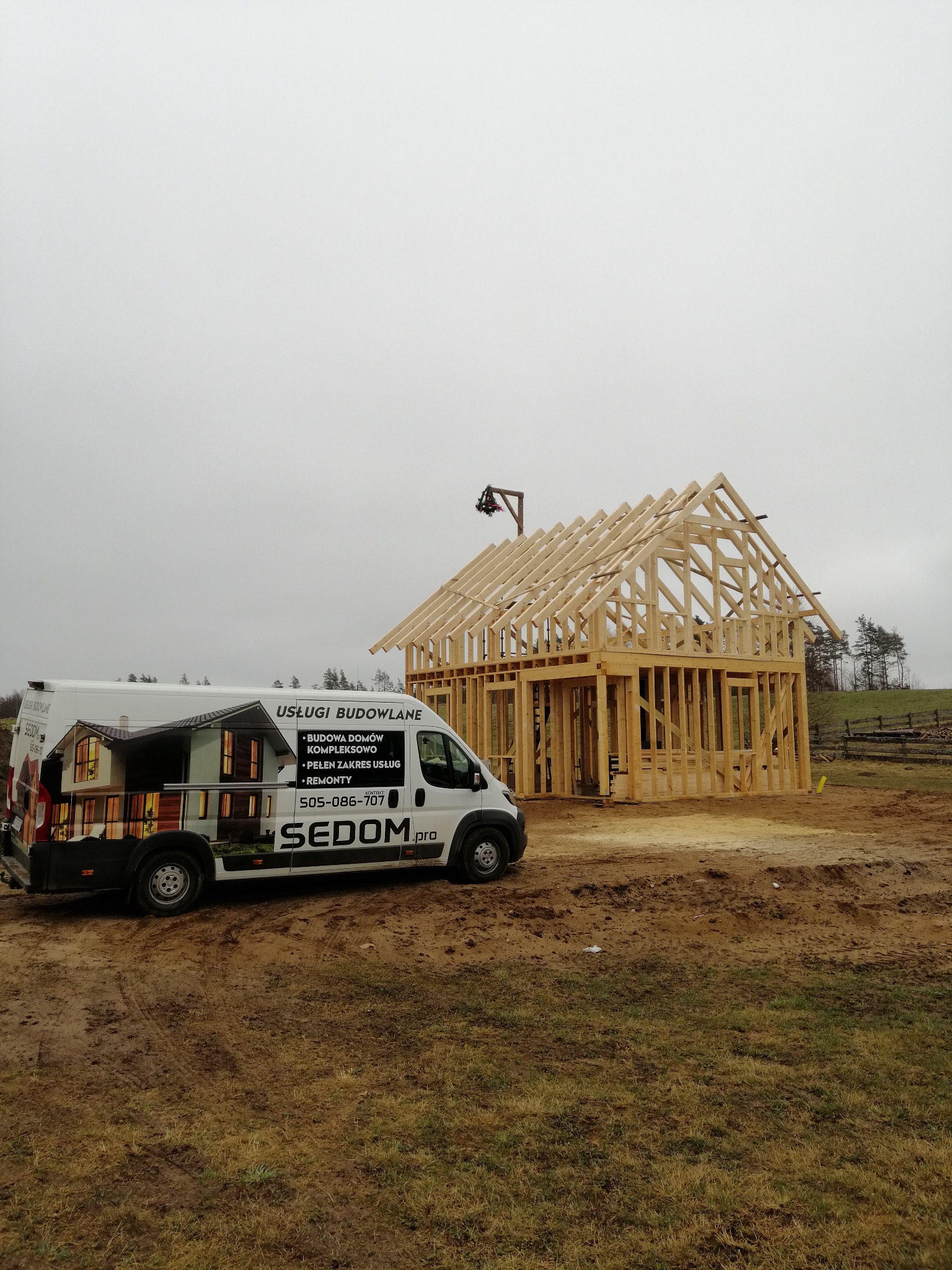 Wczesny etap budowy domu szkieletowego z widoczną konstrukcją drewnianą i zaparkowanym obok samochodem dostawczym z reklamą firmy budowlanej na tle pochmurnego nieba.