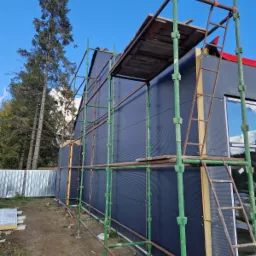 Budynek w trakcie wykończenia elewacji z użyciem rusztowania, widoczne ocieplenie i czarna fasada, wysokie drzewa w tle.