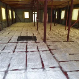 Wnętrze budynku w trakcie ocieplania: biała pianka izolacyjna naniesiona między metalowe profile podłogowe, żółta izolacja widoczna na ścianach pod oknami.