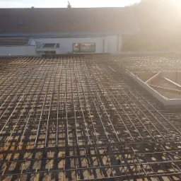 Siatka zbrojeniowa ułożona na betonowej płycie stropowej, z widoczną drewnianą szalunką otworu, w tle budynek mieszkalny w jasnym słońcu.