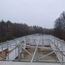 Konstrukcja stalowa dachu w trakcie montażu na murach z bloczków, widok na las w pochmurny dzień.