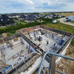 Budowa domu jednorodzinnego: widok z góry na fundamenty i ściany parteru w trakcie wznoszenia. Widoczne szalunki, zbrojenia oraz pracownicy budowlani w trakcie pracy, a w tle zabudowa jednorodzinna...