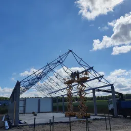 Montaż stalowej konstrukcji hali przemysłowej przez ekipę budowlaną z użyciem podnośnika nożycowego, na tle błękitnego nieba z chmurami.