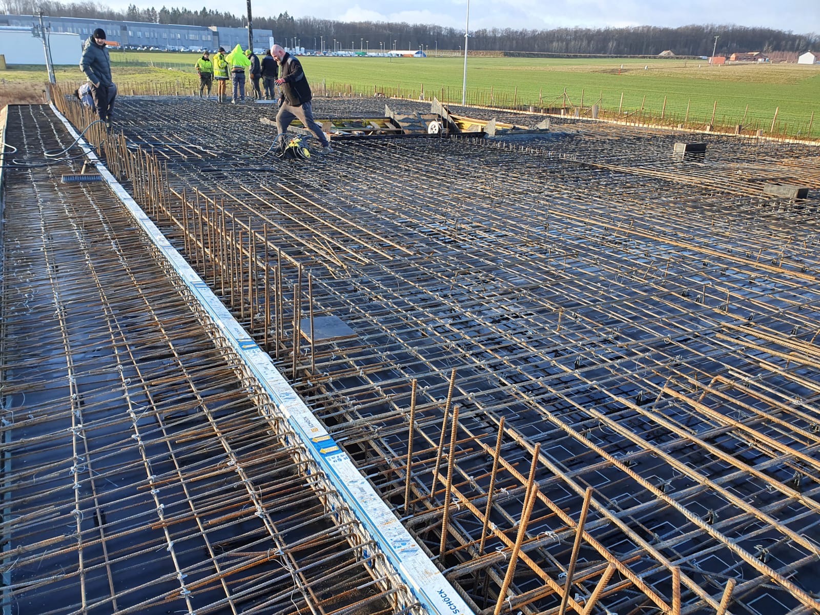 Siatka zbrojeniowa ułożona na czarnej folii, przygotowana do zalania betonem, widoczni pracownicy budowlani w tle, teren budowy na obrzeżach miasta.