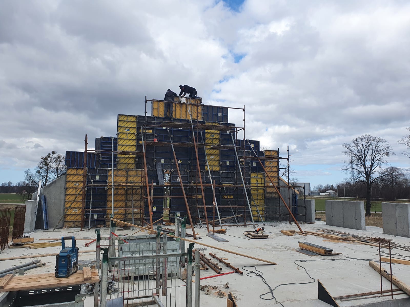 Konstrukcja szalunkowa na budowie z pracownikami na rusztowaniu, widoczne elementy zbrojenia i sprzęt budowlany na betonowej płycie fundamentowej.