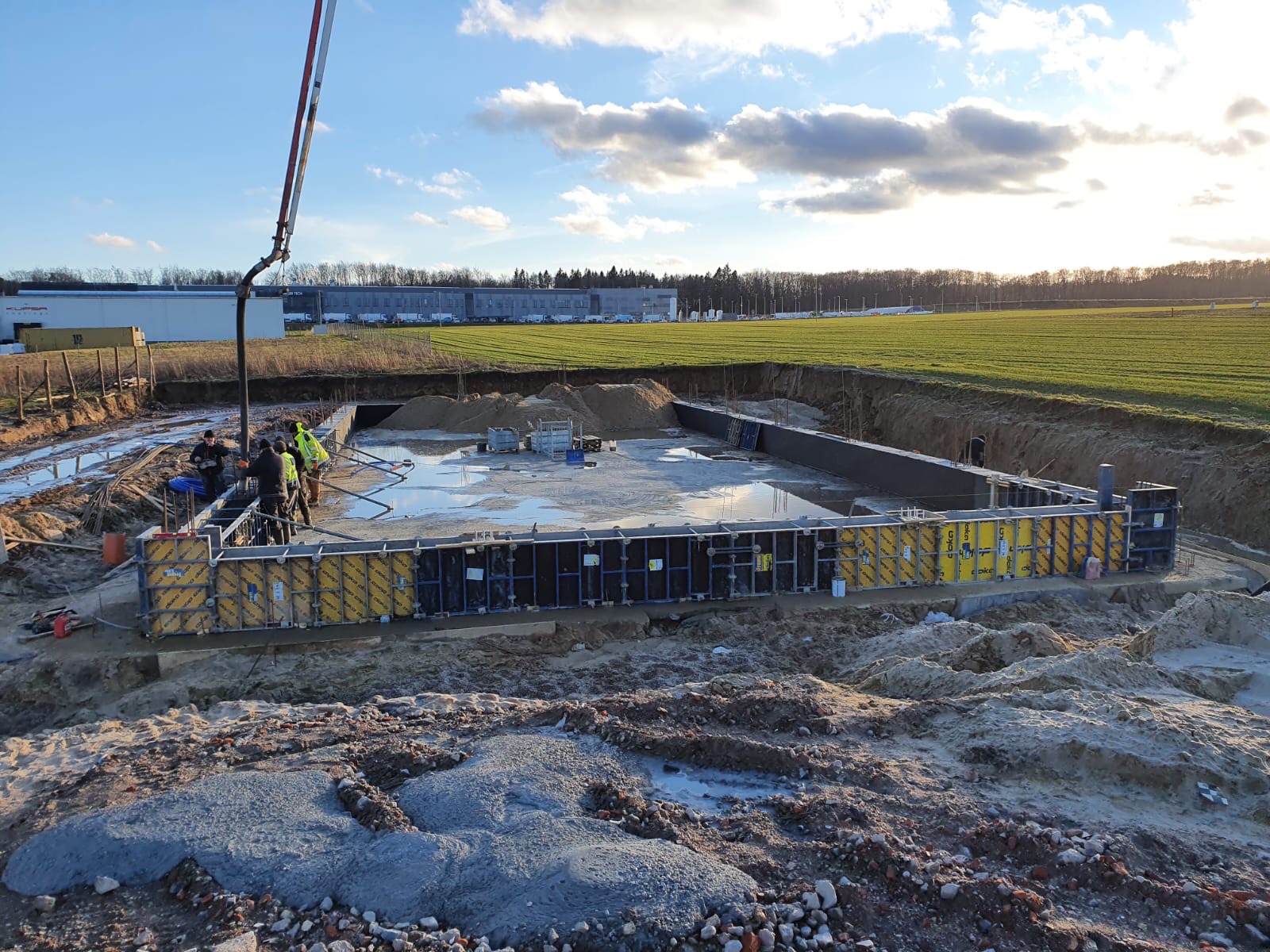 Wylewanie betonu do szalunków fundamentowych, widoczni pracownicy w odblaskowych kamizelkach i ramię pompy do betonu. W tle budynki przemysłowe i pola uprawne pod błękitnym niebem z chmurami.