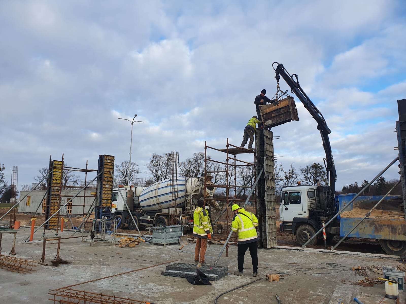 Montaż szalunków pionowych na budowie, z użyciem dźwigu i rusztowań, na tle betoniarki i pochmurnego nieba.