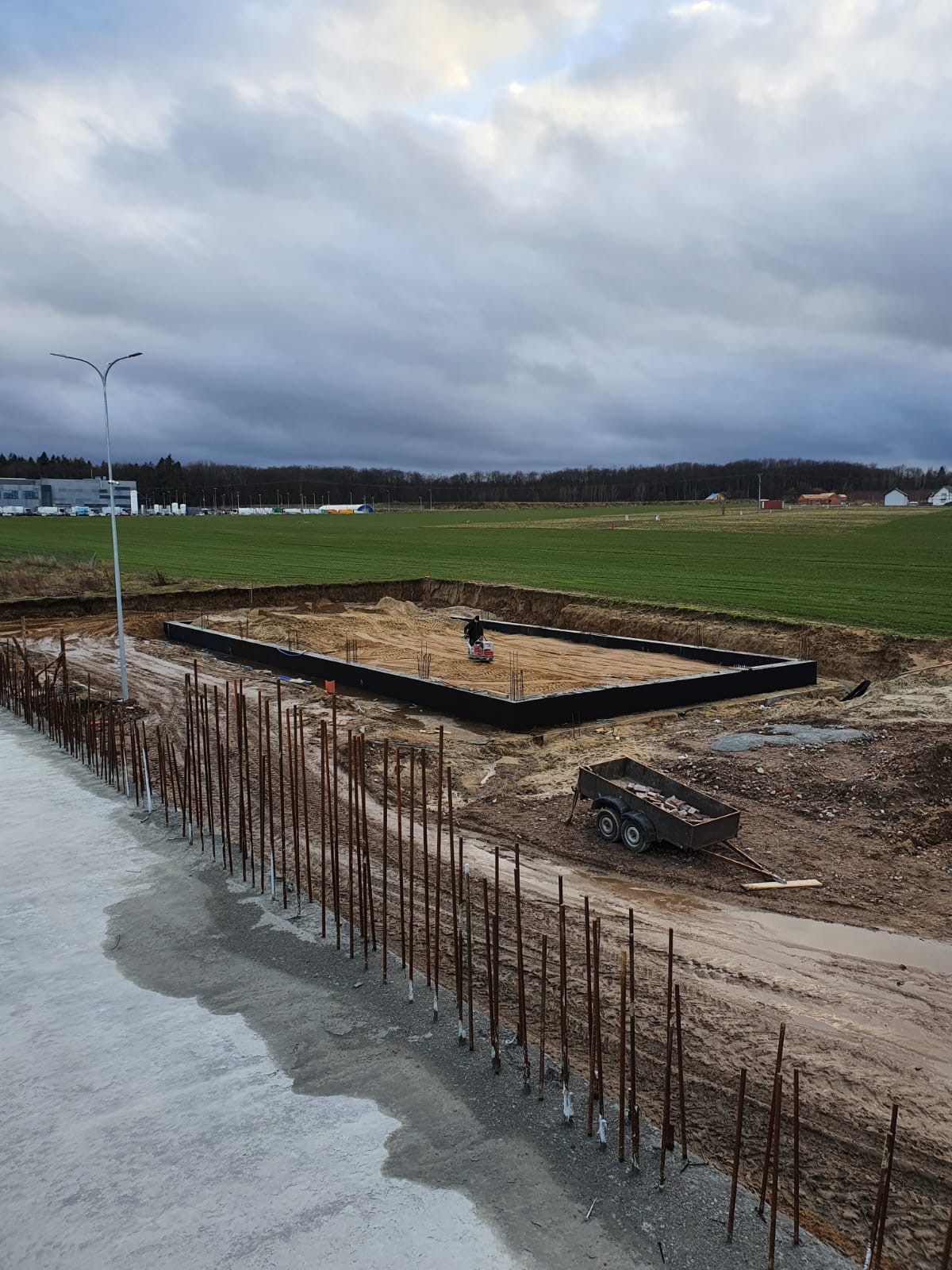 Przygotowanie terenu pod budowę domu: widok na wykopane fundamenty z czarną izolacją, zagęszczarkę w akcji, przyczepkę z cegłami i wystające pręty zbrojeniowe na pierwszym planie, w tle zielone...