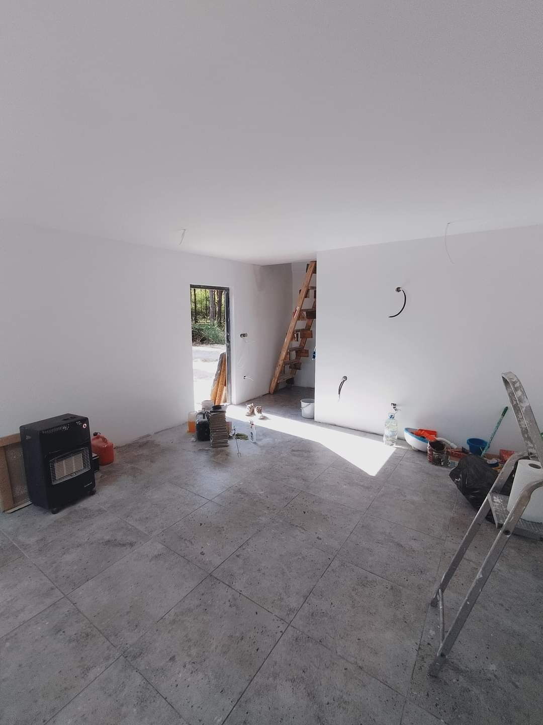 Wnętrze pokoju w trakcie remontu z nową szarą podłogą wyłożoną płytkami, widoczne narzędzia, drabina i grzejnik, ściany pomalowane na biało, w tle schody drewniane.