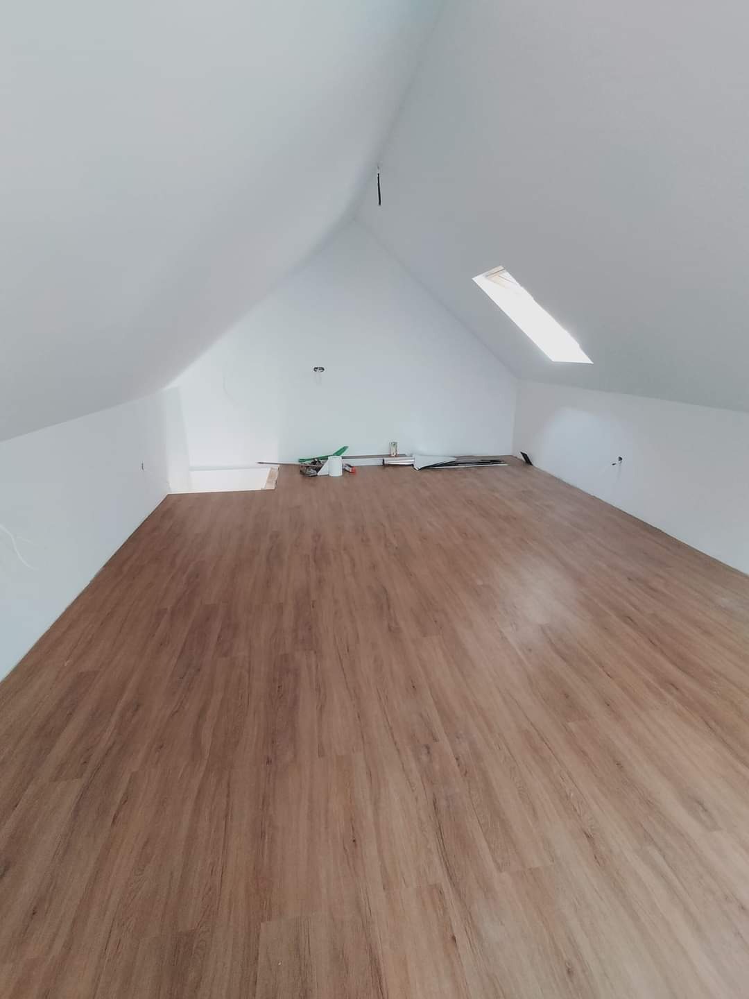 Poddasze z ukończonym układaniem paneli podłogowych w odcieniach drewna, widoczne narzędzia i materiały budowlane przy ścianie, okno dachowe wpuszczające światło.