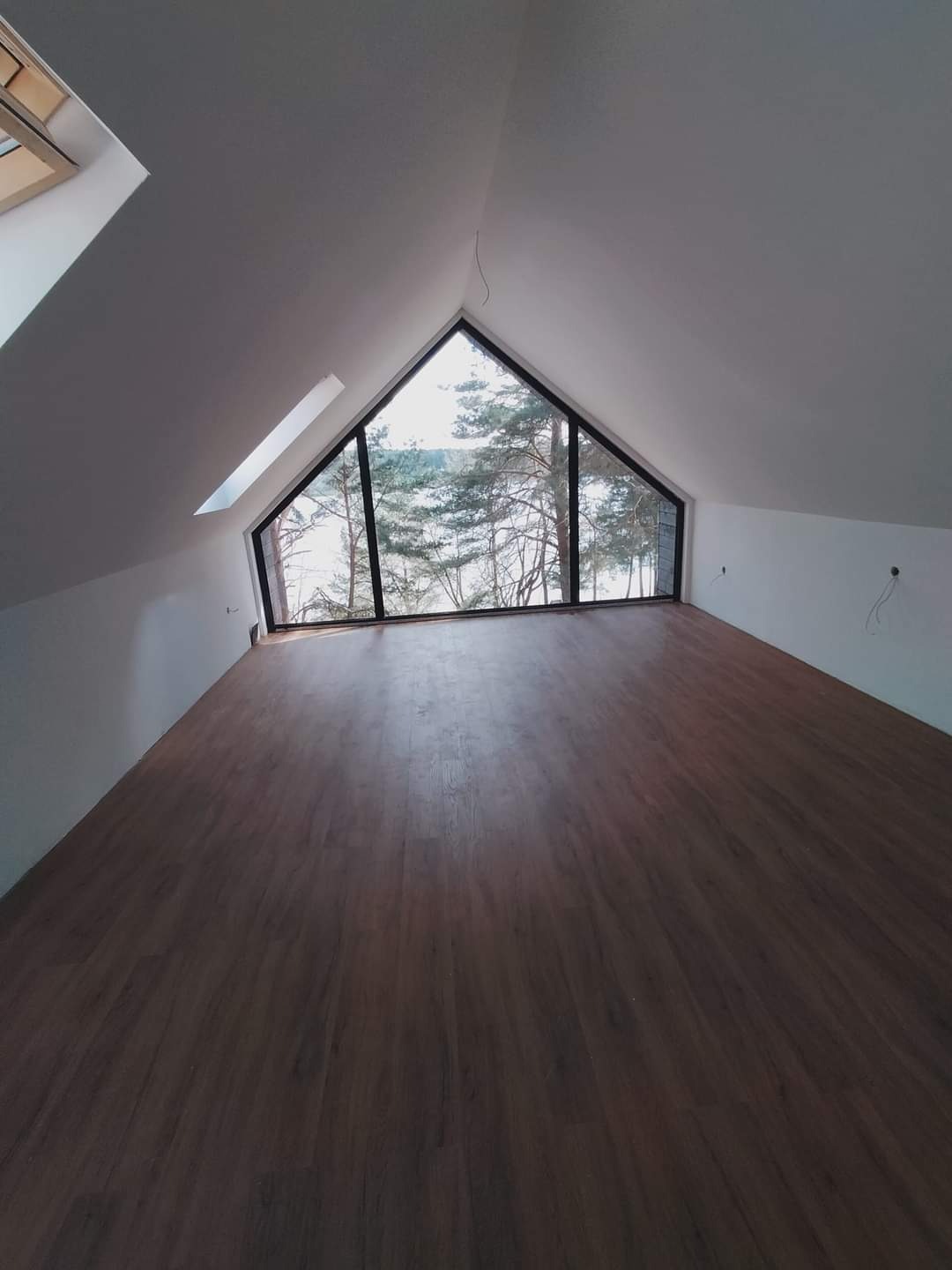 Poddasze z dużym trójkątnym oknem z widokiem na jezioro i las, świeżo położone ciemne panele podłogowe, białe ściany, okno dachowe po lewej stronie.